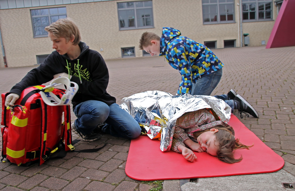 Acht Schüler stehen mit verbundenen Köpfen und Händen vor einer Schultafel. Darauf steht unter anderem: „Hautverletzung. Was tun?“